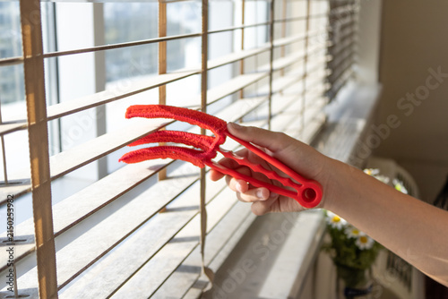 Obraz na płótnie Hand cleaning white venetian window blinds with a red slotted duster brush at a sunlit window, housekeeping closeup