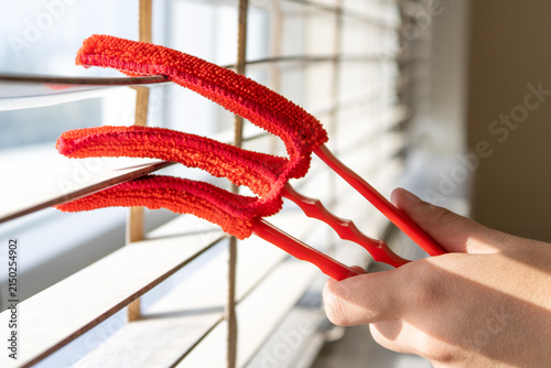 Fotografie Hand cleaning white venetian window blinds with a red slotted duster brush at a sunlit window, housekeeping closeup