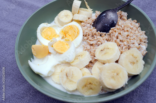 Healthy fitness breakfast bowl with oatmeal, sliced bananas, boiled eggs, and milk on a grey textile background. High protein morning meal for healthy lifestyle and balanced diet.