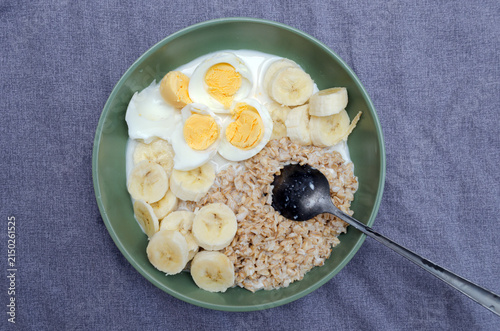 Top view of a healthy fitness breakfast bowl with oatmeal, sliced bananas, boiled eggs, and milk in a green ceramic bowl on a grey textile background.