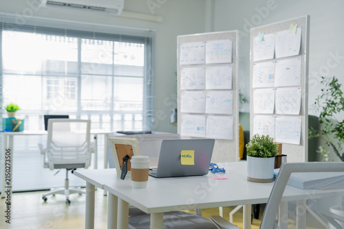 An empty white desk holds a closed laptop with a work sticky note, coffee cup, and picture frame, set against a bright office backdrop with wall mounted planning charts a clean, minimalist workspace s