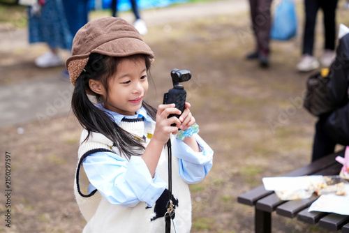 Cute Little Asian Girl Holding a Small Pocket holding a compact gimbal camera to record video or take photos while standing outdoors in a park.