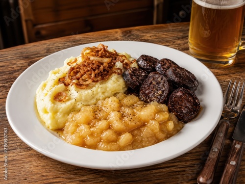 Himmel und Erde im deutschen Restaurant – deutscher Klassiker mit Kartoffelpüree, Apfelkompott, Blutwurst und Röstzwiebeln, deftig angerichtet auf weißem Teller mit Bier.