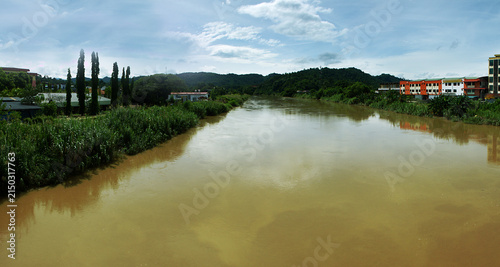 An artificial drainage canal with muddy water during the rainy season, a straightened river amidst tropical forest. A village on the shore and hills on the horizon. Indonesia.