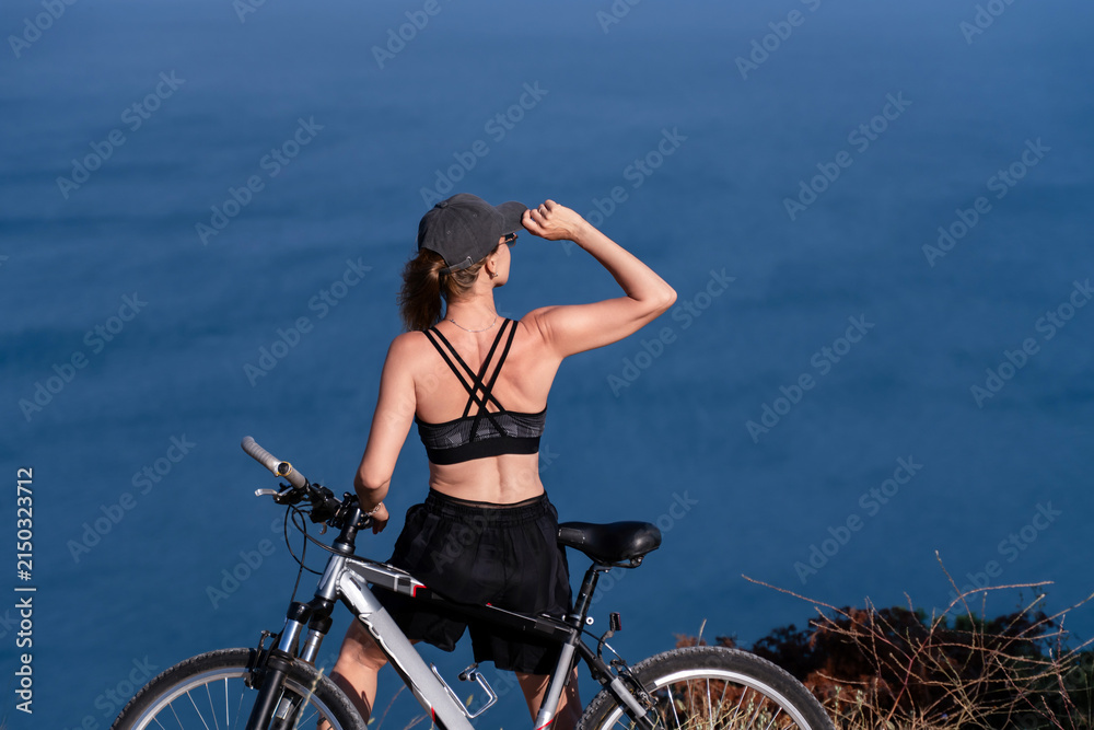 Fototapeta premium Bicycle, cyclist, ocean, woman with mountain bike standing on cliff top looking at sea during sunny day for outdoor fitness inspiration