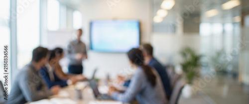 A business meeting in a modern office setting, with a blurred group of professionals listening to a presentation on a screen. Collaboration and teamwork in focus.