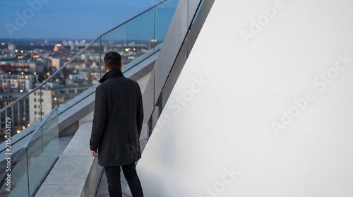 Man standing on rooftop terrace overlooking city at night. Business person looking at urban skyline. Concept of success, future, leadership and ambition. Modern architecture exterior.