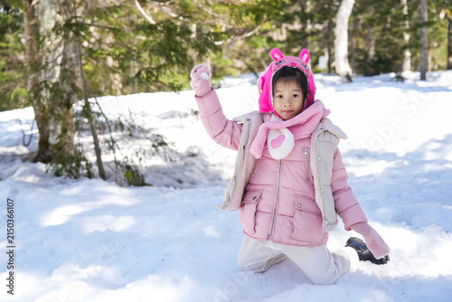 Little Asian Girl wearing a pink winter jacket winter clothes kneeling and Throwing a Snowball in pine Forest.