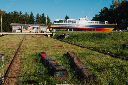 Canvastavla Old passenger boat standing in grassy dry dock with rails, wooden beams, workshop and clear blue sky