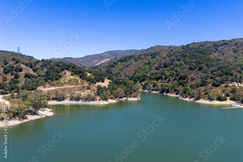 Lexington Reservoir water reflecting clear blue sky, surrounded by green hills with winding roads and scattered trees under a bright summer sun