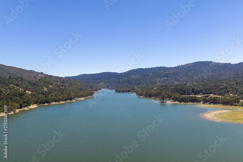 Lexington Reservoir water body stretching through the forested hills of Los Gatos, California, showing a clear blue sky and evidence of low water levels along the dry shoreline