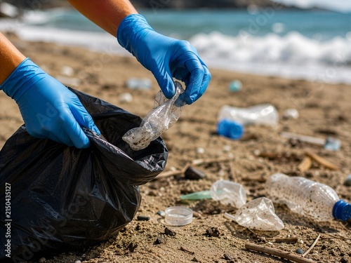 Volunteer in gloves picking up plastic bottles and trash from a beach into a garbage bag, concept of ocean pollution, beach cleanup and environmental protection