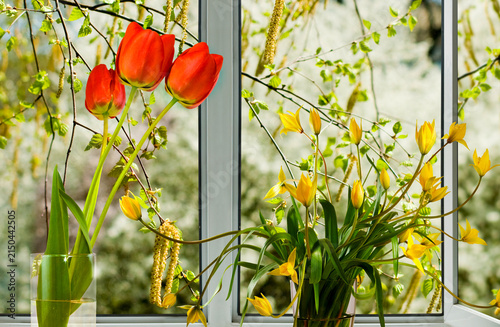 Flowers are arranged in vases by a window with greenery in the background on a sunny day