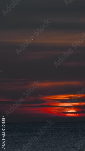 Airplane flying through dramatic orange and red clouds during sunset over the calm ocean, vertical shot, wide angle, real time