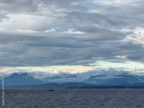 coastal mountains of southwestern Canada at dusk with sunset colors on distant clouds over land and a layer of gray clouds over the ocean, with a fishing boat looking very small in the vast landscape