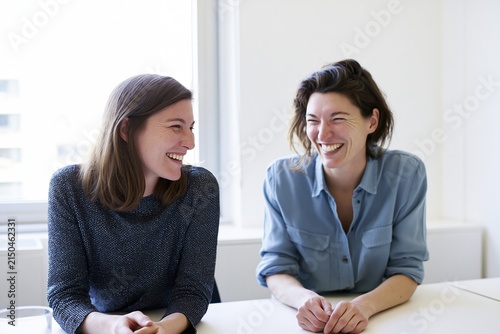 two adult coworkers sharing a genuine laugh in a bright minimal office, seated at a plain table, hands below frame