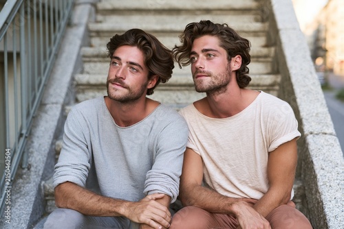 two young adult men sitting shoulder to shoulder on apartment stairs, relaxed casual clothes, looking into the distance