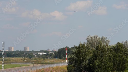 Airplane flying high in the sky above airport runway with distant city buildings under a bright blue sky on a sunny day, wide static shot