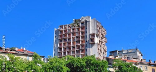 Cuadro en lienzo An abandoned high-rise building in Rijeka, Croatia, to be renovated for affordable public housing, standing above lush green trees under a bright blue sky, promoting modern urban revitalization
