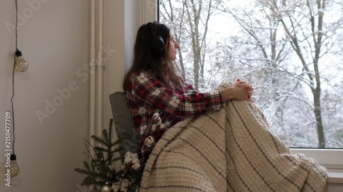 Young woman in pajamas and headphones sits by a window wrapped in a cozy blanket, listening to music and peacefully watching snow falling outside.