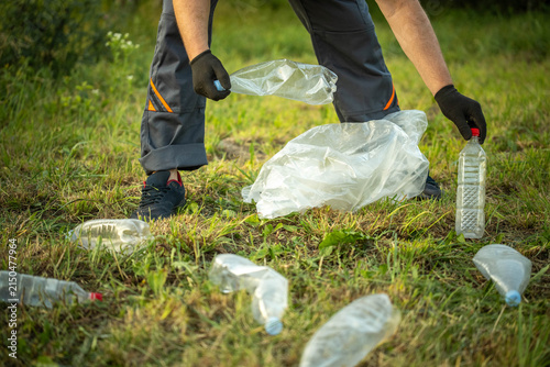 A janitor wearing gloves picks up plastic bottles and wraps them in a bag on the grass. They are participating in a clean-up event to help the environment in a local park
