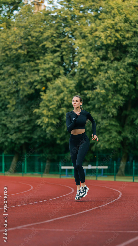 Fototapeta premium Young woman running on a red track with green trees in the background