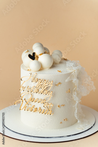 Elegant white birthday cake with lace decoration and golden topper with Victoria name on the beige background