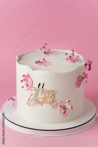 Elegant white birthday cake with pink blossoms and gold Happy Birthday topper on the pink background