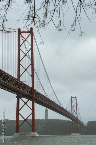 View of the 25 de Abril Bridge in Lisbon stretching across the Tagus River on a cloudy day, with soft mist surrounding the iconic red suspension structure and calm urban waterfront scenery.