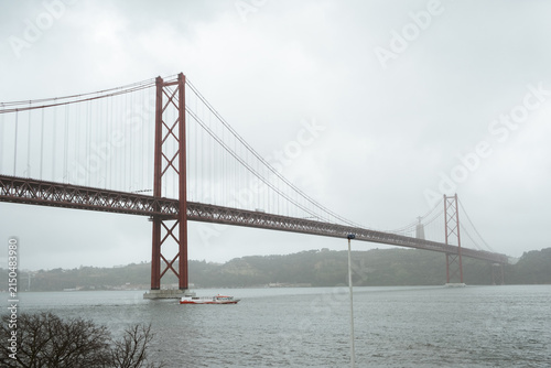 View of the 25 de Abril Bridge in Lisbon stretching across the Tagus River on a cloudy day, with soft mist surrounding the iconic red suspension structure and calm urban waterfront scenery.