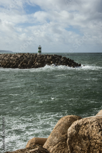 Green and white lighthouse standing on rocky harbor barrier as Atlantic Ocean waves crash along the coast in Portugal beneath bright cloudy sky and dramatic seaside scenery.