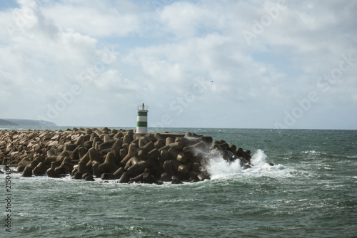 Green and white lighthouse standing on rocky harbor barrier as Atlantic Ocean waves crash along the coast in Portugal beneath bright cloudy sky and dramatic seaside scenery.