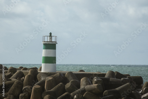 Green and white lighthouse standing on rocky harbor barrier as Atlantic Ocean waves crash along the coast in Portugal beneath bright cloudy sky and dramatic seaside scenery.
