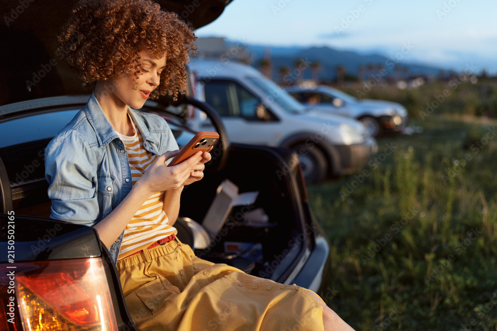Obraz premium Cheerful woman outdoors, relaxed lifestyle portrait, sitting at car trunk, checking phone, warm sunlight, denim jacket, striped tee, meadow scenery, gentle breeze, natural colors