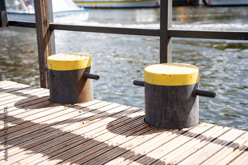 Two black and yellow mooring bollards on a wooden pier deck next to the water with a blurry boat in the background.