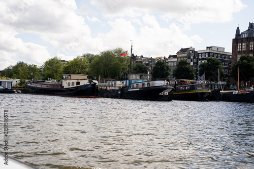Traditional houseboats and historical barges moored along the canal in Amsterdam under a cloudy summer sky.