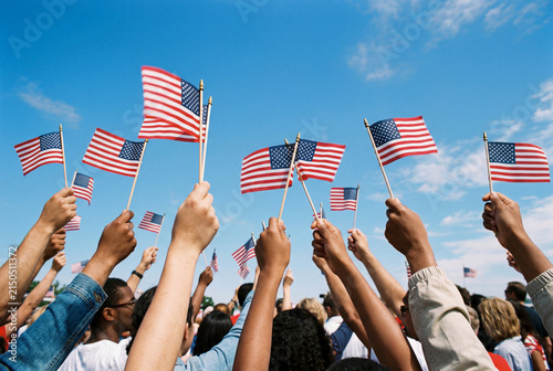 Diverse crowd raising hands with american flags against blue sky