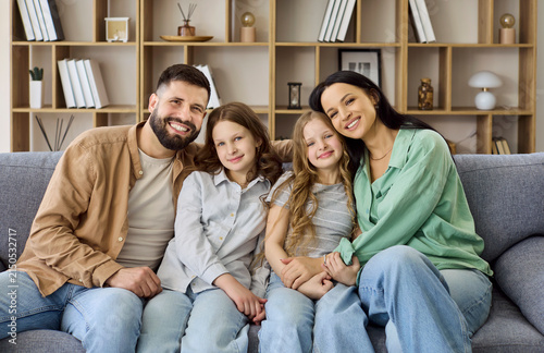 Family parents daughters portrait smiling together at home. Mother and father hug their daughters on a cozy sofa in a living room, perfect for lifestyle ads. Ideal concept for family unity