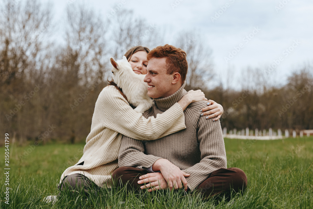 Obraz premium An affectionate couple sits on the grass in a sunny field, sharing a warm hug with a friendly goat kid between them. A candid lifestyle moment capturing connection, nature, and rural life.