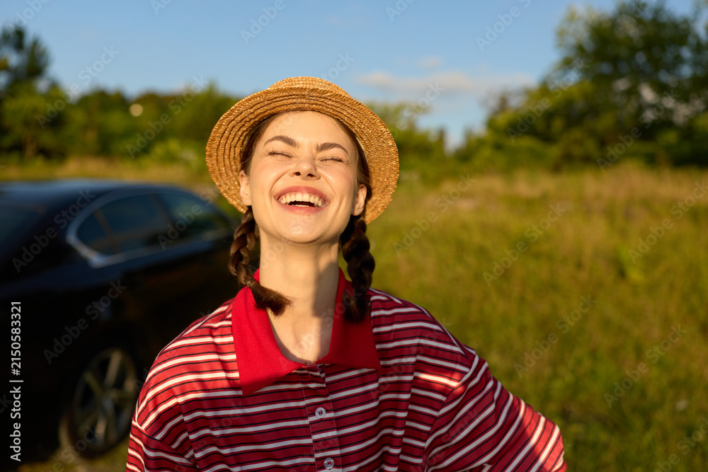 Obraz premium Young woman in a striped red shirt and straw hat smiles with eyes closed outdoors, standing in a sunny field. Natural light, lifestyle concept focused on happiness and leisure.