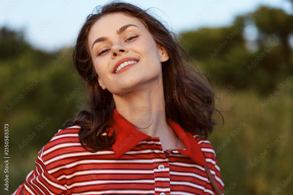 Obraz premium A smiling young woman in a striped polo shirt enjoys the warm sunlight outdoors. Lifestyle portrait of a happy girl in nature, concept of freedom, youth, joy and wellbeing.