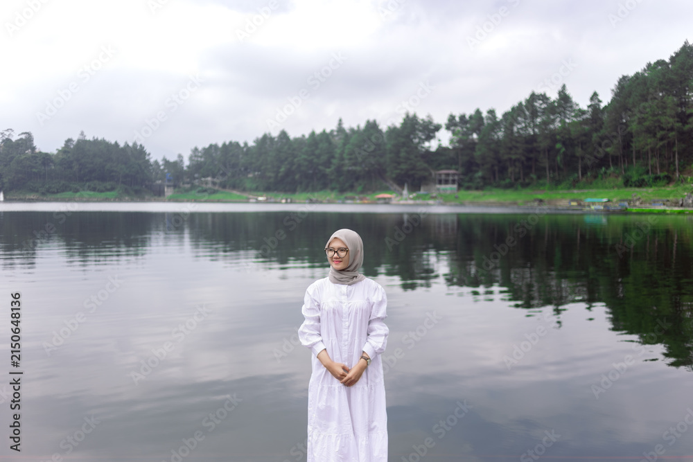 Fototapeta premium Portrait of a Muslim woman wearing a white hijab on the bank of a river. A young woman meets a summer daylight by the water.