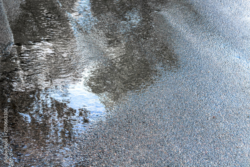 reflections of green trees and blue sky in a puddle on asphalt road.
