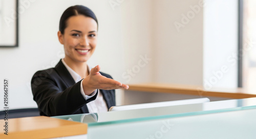 Elegant female receptionist offering an open hand welcome at a bright modern desk perfect for a friendly hospitality service concept