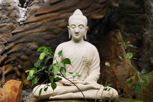 White Buddha statue in sitting meditation posture surrounded by natural rocks and Bodhi tree leaves, symbol of peace and enlightenment.