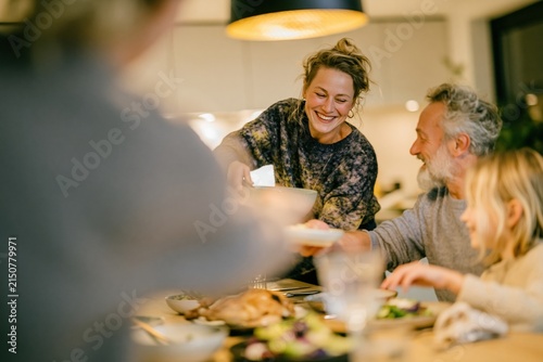 Smiling woman serving food to her multi-generational family at a wooden dining table, warm and hospitable atmosphere for home cooking and togetherness campaigns