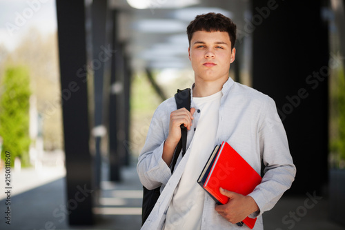 Young man stands outside a building with books and a backpack during daytime in a modern area