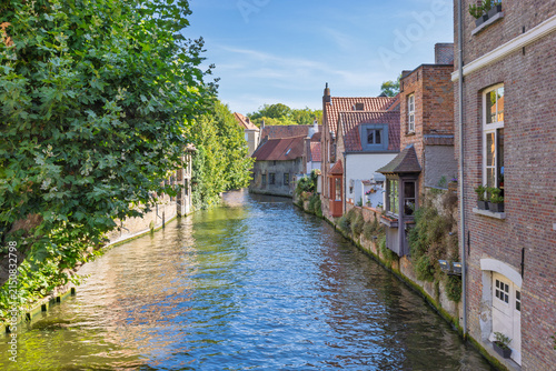 Bruges town in Belgium, Europe. Charming canal scene with historic buildings and lush green trees along the tranquil waterway under a bright blue sky on a sunny day