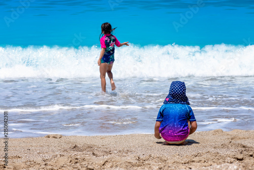 girls palying on the beach wearing sun protective clothes, two sisters enjoying hot summer day on the beach 