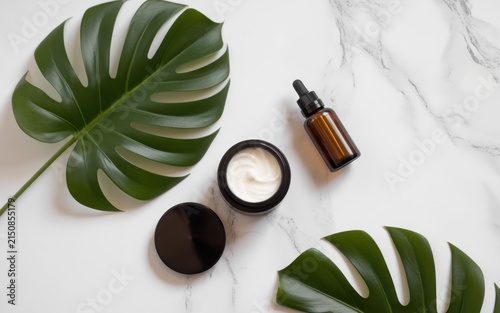 Skincare beauty products with organic face cream in a jar and brown glass serum bottle on a white marble surface decorated with large green monstera tropical leaves